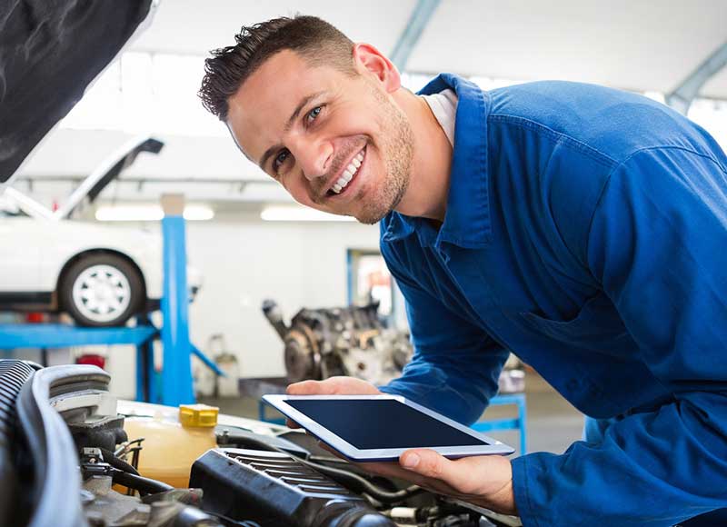 service mechanic with ipad standing over car engine, smiling at camera